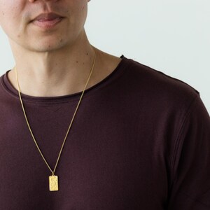 Pu&ograve; includere: Una persona indossa una maglietta color granata con una collana a catena dorata e un ciondolo d'oro. Il ciondolo &egrave; di forma rettangolare con un design testurizzato.