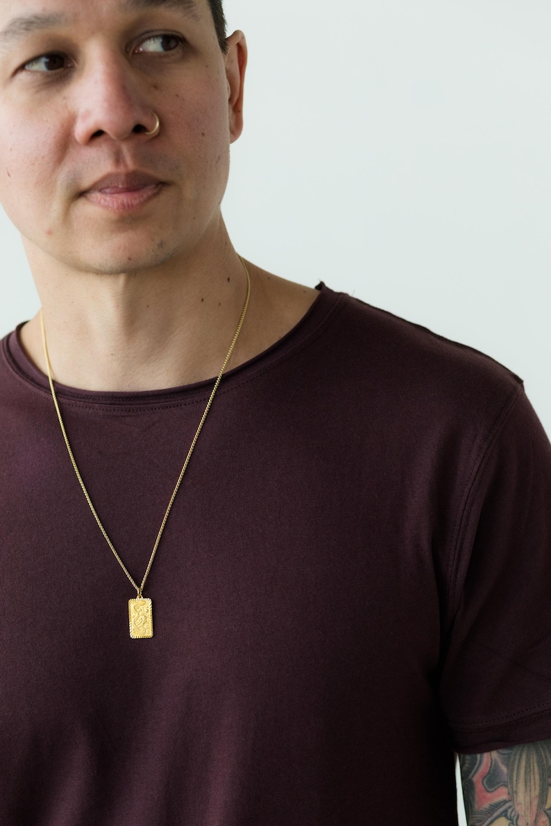 Pu&ograve; includere: Una persona indossa una maglietta color granata con una collana a catena dorata e un ciondolo d'oro. Il ciondolo &egrave; di forma rettangolare con un design testurizzato.