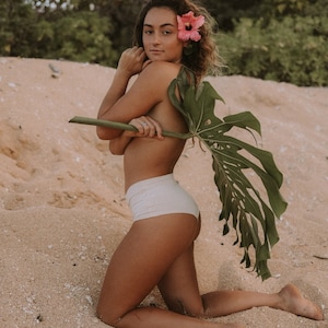 May include: A woman wearing a white bikini bottom poses on a sandy beach. She is holding a large green leaf behind her back and has a pink flower in her hair.