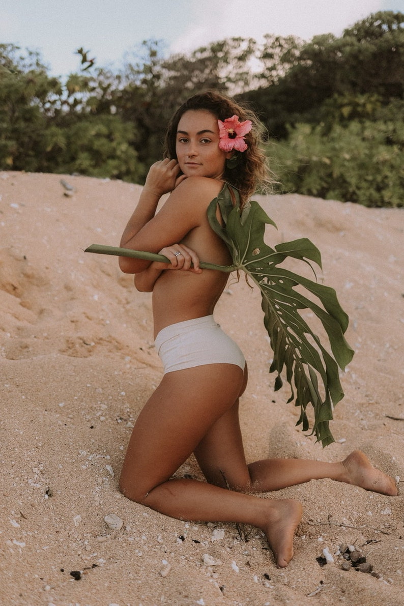 May include: A woman wearing a white bikini bottom poses on a sandy beach. She is holding a large green leaf behind her back and has a pink flower in her hair.
