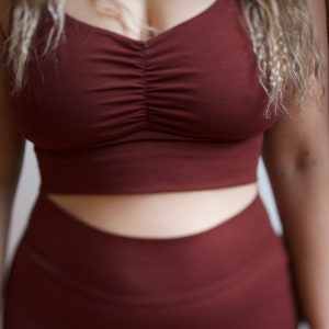 May include: Two women wearing matching maroon sports bras and leggings. The sports bra has a scoop neck and a gathered detail in the front.