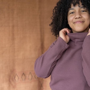 May include: A woman with curly dark hair is wearing a mauve turtleneck sweater. She is smiling and looking at the camera.