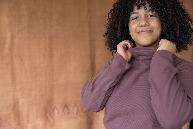 May include: A woman with curly dark hair is wearing a mauve turtleneck sweater. She is smiling and looking at the camera.