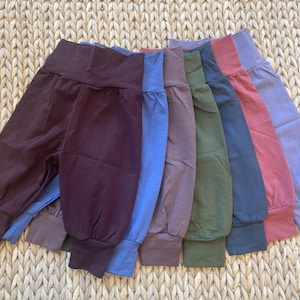 May include: A collection of seven pairs of solid-coloured trousers with high, wide waistbands and cuffed ankles. The trousers are arranged in a fan-like display, featuring colours including dark brown, blue, mauve, olive green, slate blue, dusty rose, and lavender.