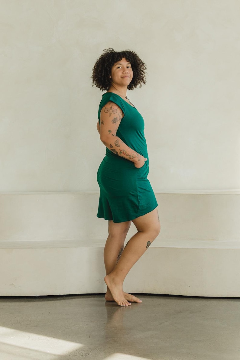 May include: A solid dark emerald green sleeveless dress featuring a scoop neckline, a relaxed fit, and side pockets. The dress falls to a length above the knee, showcasing a simple, clean silhouette with wide shoulder straps and a straight hemline.