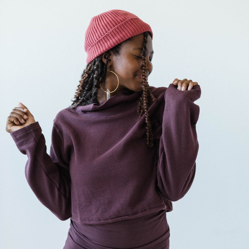 May include: A woman wearing a maroon ribbed turtleneck sweater with long sleeves and a pink beanie. She is wearing large hoop earrings and has long, dark hair.