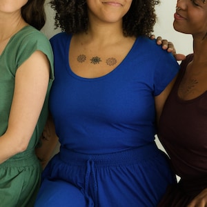 May include: Three people wearing scoop-neck tops and matching bottoms in shades of green, blue, and burgundy. The person in the center has a small tattoo on their chest. The clothing appears to be made of a soft, comfortable material.