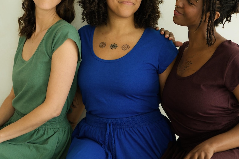 May include: Three people wearing scoop-neck tops and matching bottoms in shades of green, blue, and burgundy. The person in the center has a small tattoo on their chest. The clothing appears to be made of a soft, comfortable material.