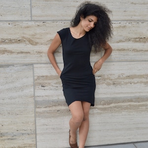 May include: A black knee-length dress with short sleeves, a rounded neckline, and side pockets. The garment features a fitted bodice that transitions into a slightly relaxed skirt, providing a simple silhouette with no additional patterns or embellishments.