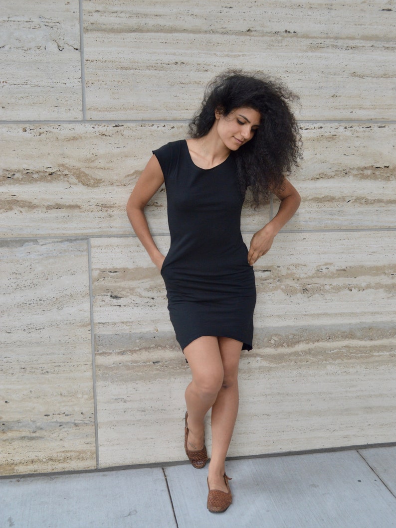 May include: A black knee-length dress with short sleeves, a rounded neckline, and side pockets. The garment features a fitted bodice that transitions into a slightly relaxed skirt, providing a simple silhouette with no additional patterns or embellishments.