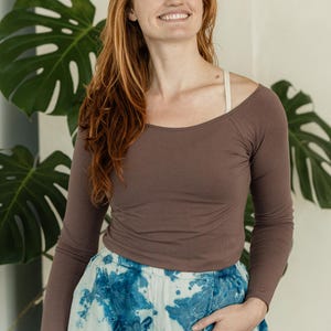 以下が含まれることがあります： 広いラウンドネックと肩に見える明るい色のストラップが特徴の茶色の長袖シャツと、ギャザー入りの伸縮性のあるウエストバンドを備えた、ゆったりとしたシルエットのライトブルーと白のタイダイ柄スカート。
