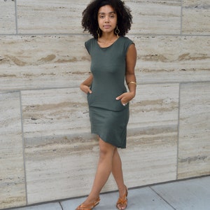 May include: A dark olive green short-sleeved dress featuring a round neckline, two side pockets, and an asymmetrical hemline. The dress is paired with gold hoop earrings, a gold bangle bracelet, and brown leather strappy sandals.