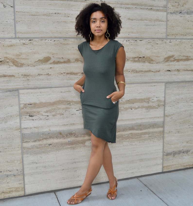 May include: A dark olive green short-sleeved dress featuring a round neckline, two side pockets, and an asymmetrical hemline. The dress is paired with gold hoop earrings, a gold bangle bracelet, and brown leather strappy sandals.