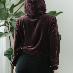 May include: A burgundy velvet hoodie with a drawstring waist and a matching hood. The model is wearing black trousers. The outfit is casual and comfortable.