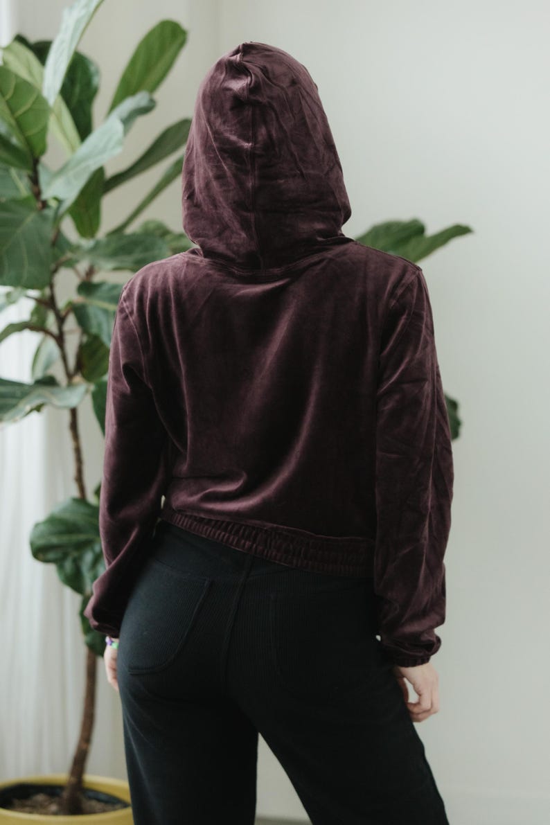 May include: A burgundy velvet hoodie with a drawstring waist and a matching hood. The model is wearing black trousers. The outfit is casual and comfortable.