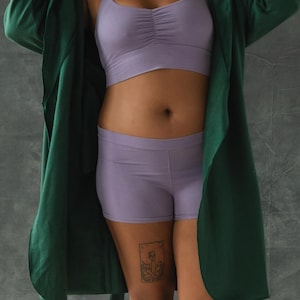 May include: A woman wearing a green satin robe over a lavender crop top and matching shorts. She has a tattoo on her thigh.
