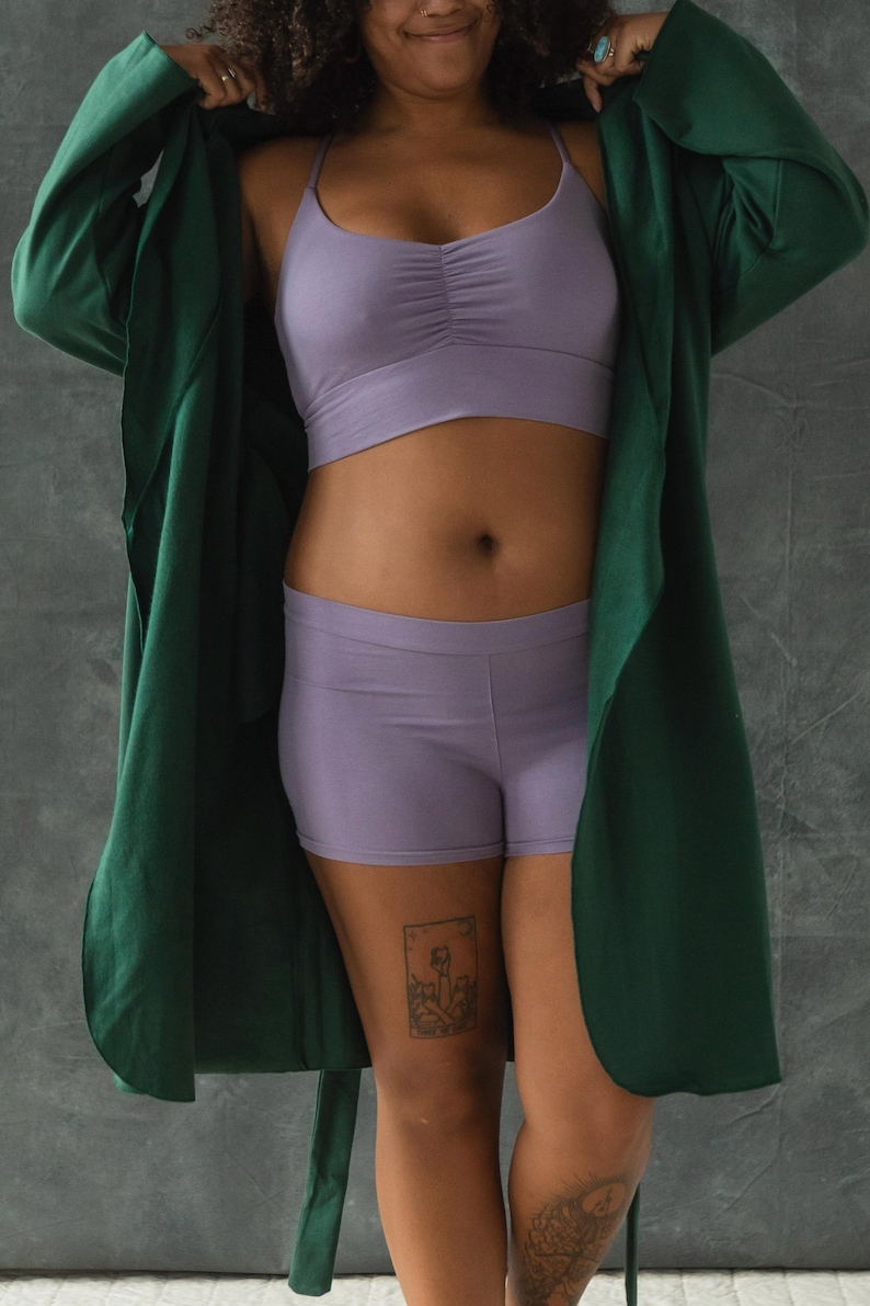 May include: A woman wearing a green satin robe over a lavender crop top and matching shorts. She has a tattoo on her thigh.