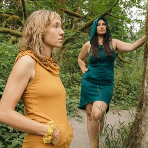 May include: Two people wearing hooded dresses stand in a forest. The person on the left is wearing a mustard yellow dress and the person on the right is wearing a teal green dress.