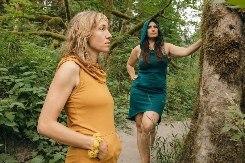 May include: Two people wearing hooded dresses stand in a forest. The person on the left is wearing a mustard yellow dress and the person on the right is wearing a teal green dress.