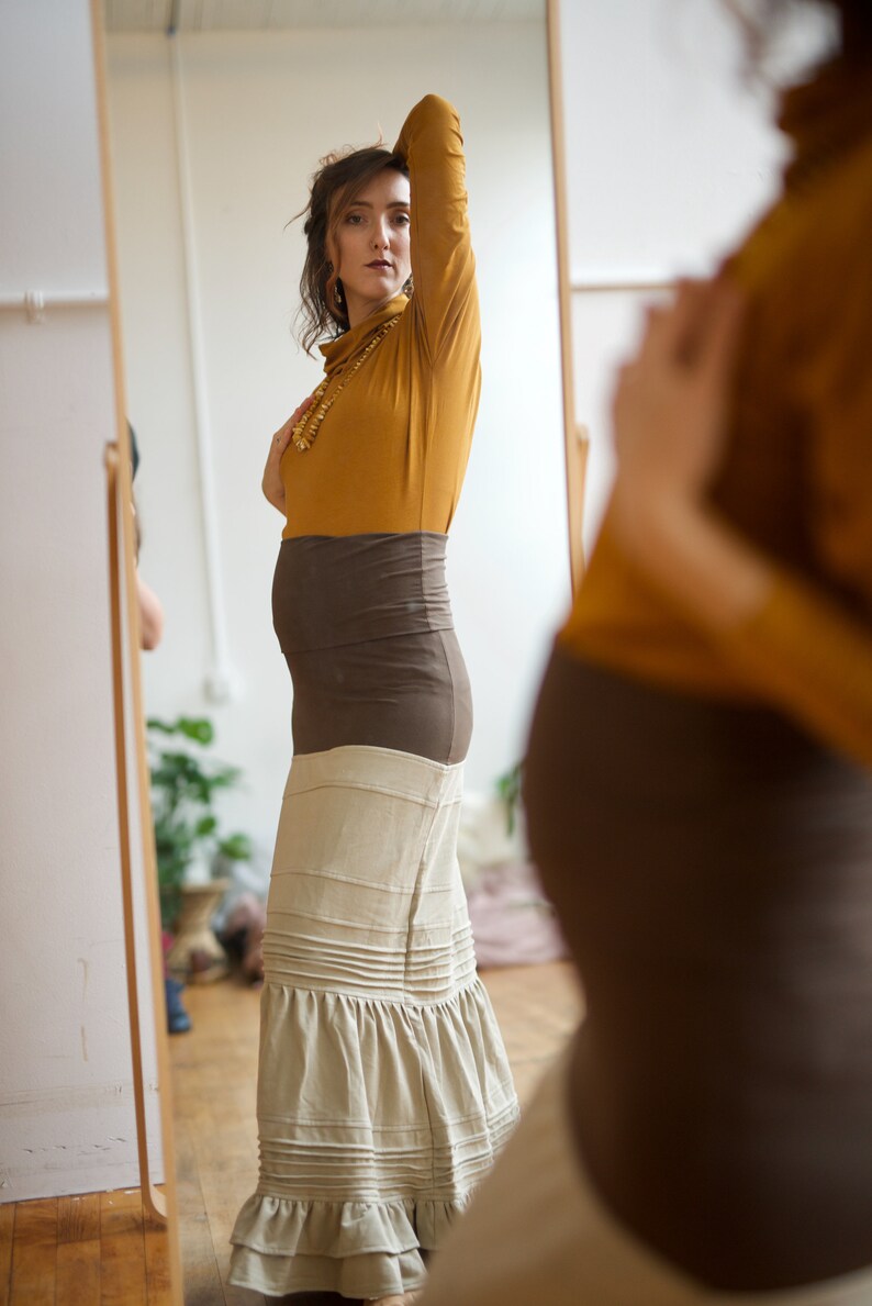 May include: A woman wearing a mustard yellow turtleneck sweater and a brown belt is standing in front of a mirror. She is wearing a long, beige skirt with a ruffled hem.