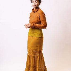 May include: A woman wearing a long, rust-coloured skirt with a ruffled hem. The skirt has a mustard yellow waistband and is paired with a rust-coloured turtleneck top.