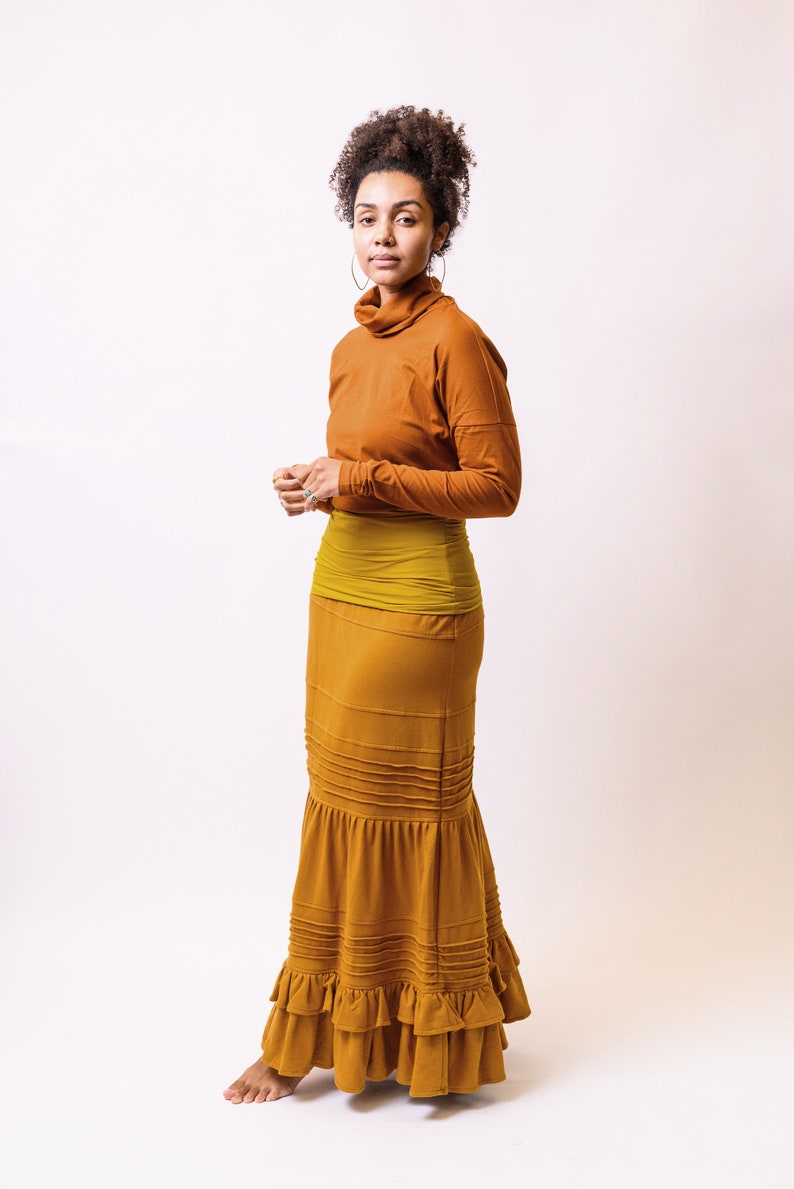 May include: A woman wearing a long, rust-coloured skirt with a ruffled hem. The skirt has a mustard yellow waistband and is paired with a rust-coloured turtleneck top.