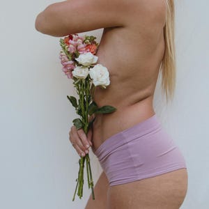 May include: A bouquet of flowers with white, pink, and orange blooms is held against a person's side. The person is wearing lavender shorts. The background is a plain white.