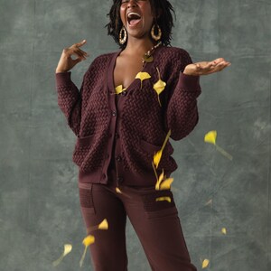 May include: A person wearing a maroon cardigan and matching pants with a textured pattern. They are laughing with their mouth open and have leaves falling around them.