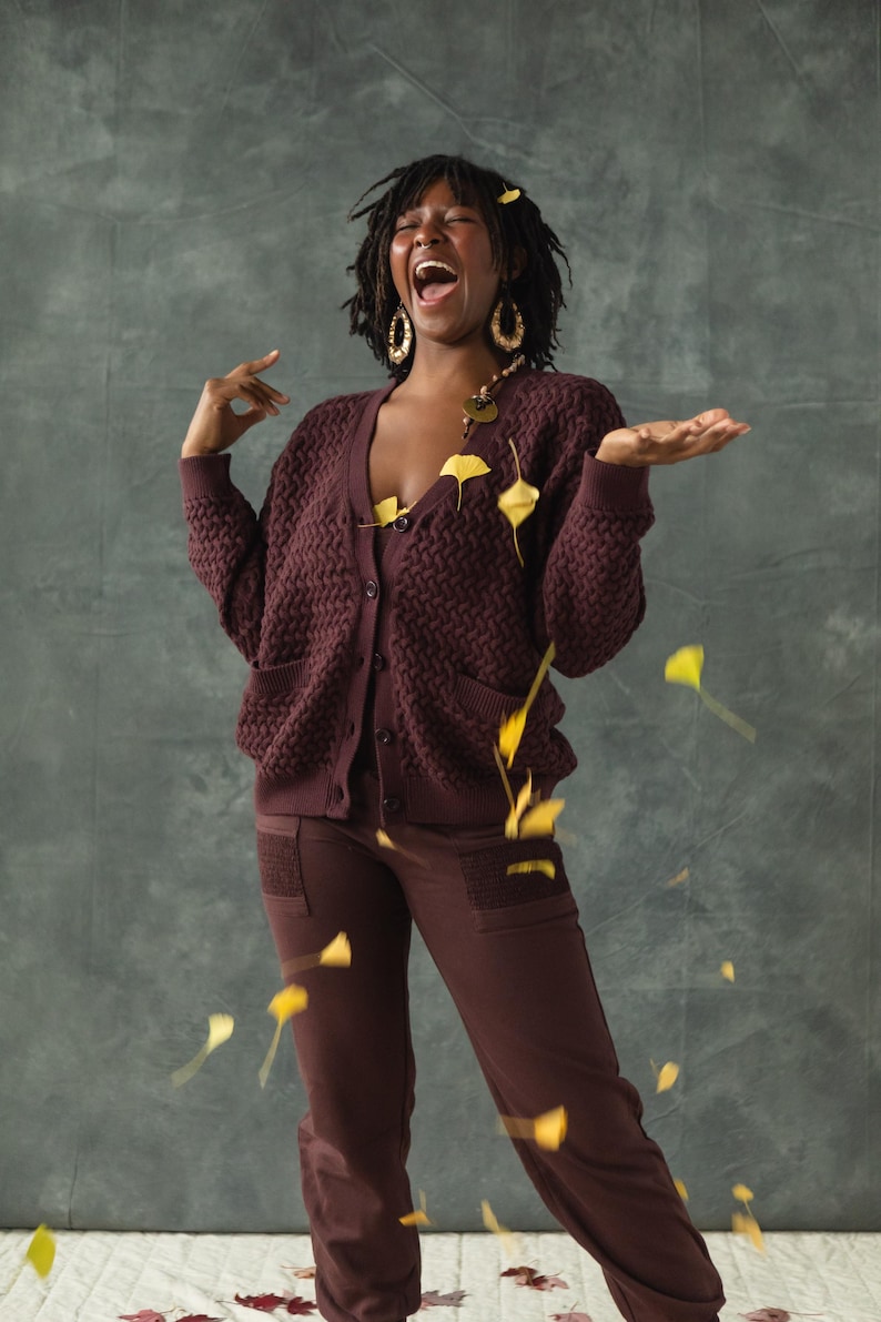 May include: A person wearing a maroon cardigan and matching pants with a textured pattern. They are laughing with their mouth open and have leaves falling around them.