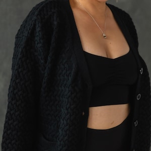 May include: A black knitted cardigan with a button closure and a textured pattern. The cardigan is worn over a black tank top and black pants.