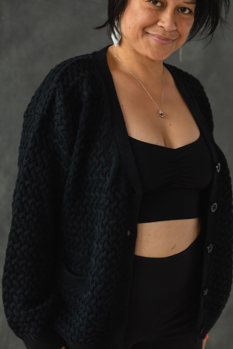 May include: A black knitted cardigan with a button closure and a textured pattern. The cardigan is worn over a black tank top and black pants.