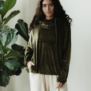 May include: An olive green velvet hoodie with a cowl neck and drawstring detail. The long-sleeved top is paired with cream-coloured trousers. The model is standing in front of a large green plant.