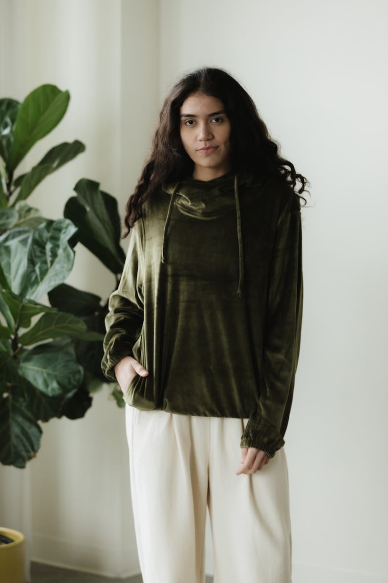 May include: An olive green velvet hoodie with a cowl neck and drawstring detail. The long-sleeved top is paired with cream-coloured trousers. The model is standing in front of a large green plant.
