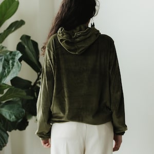 May include: An olive green hoodie with a drawstring hood and long sleeves. The hoodie is made of a soft, velvety material. The person is wearing cream-coloured trousers. The background is a plain white wall with a large green plant.
