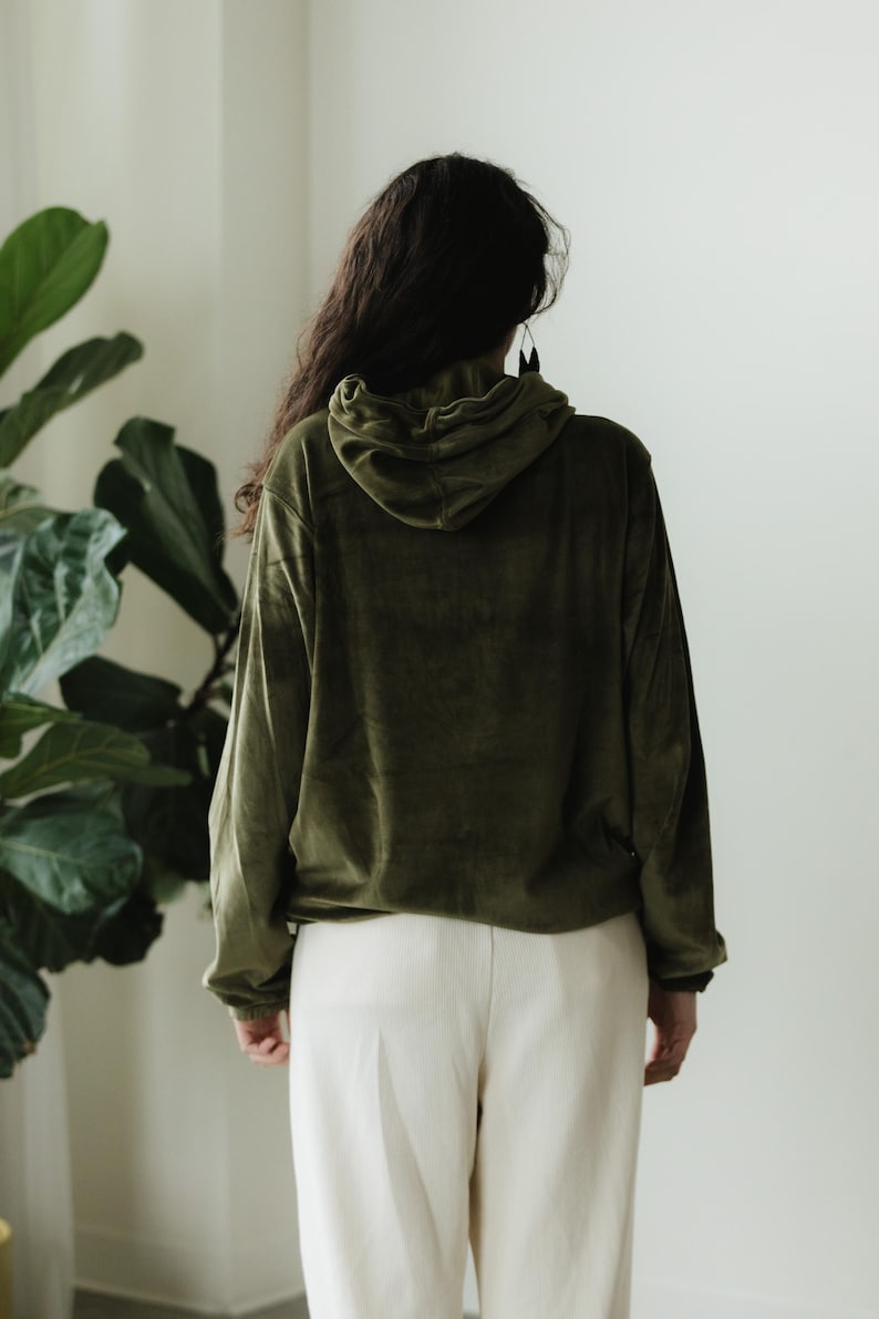 May include: An olive green hoodie with a drawstring hood and long sleeves. The hoodie is made of a soft, velvety material. The person is wearing cream-coloured trousers. The background is a plain white wall with a large green plant.