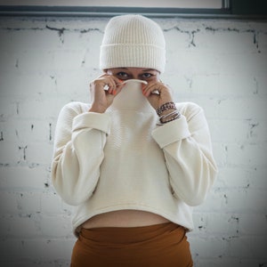 May include: A white knit sweater with a ribbed texture, worn with a white beanie and a brown skirt.