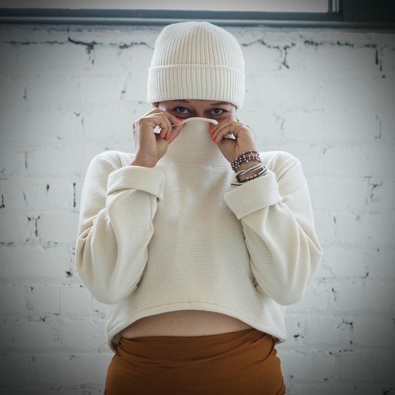 May include: A white knit sweater with a ribbed texture, worn with a white beanie and a brown skirt.