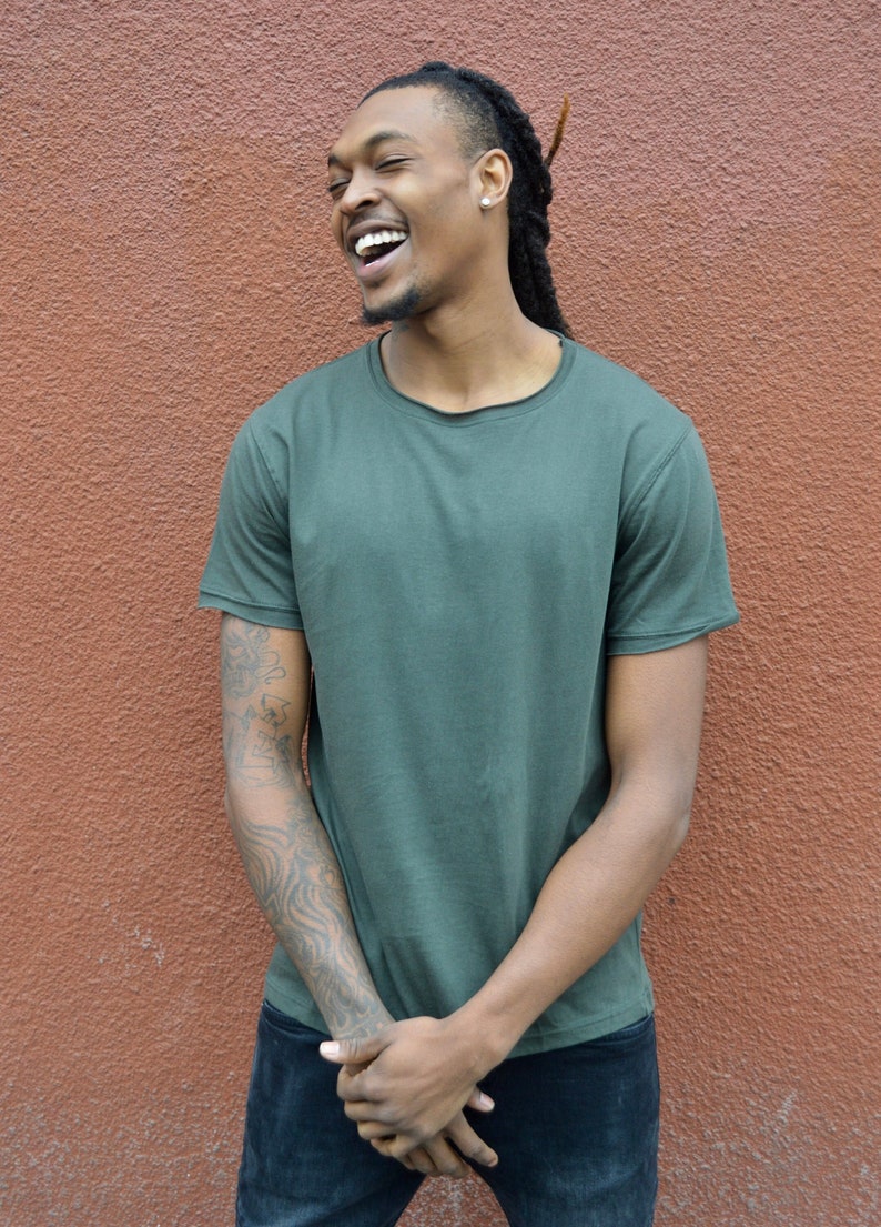 Pu&ograve; includere: Un uomo con i dreadlocks indossa una maglietta verde scuro e jeans blu. Sta sorridendo e guardando a sinistra. Ha un tatuaggio sul braccio sinistro.