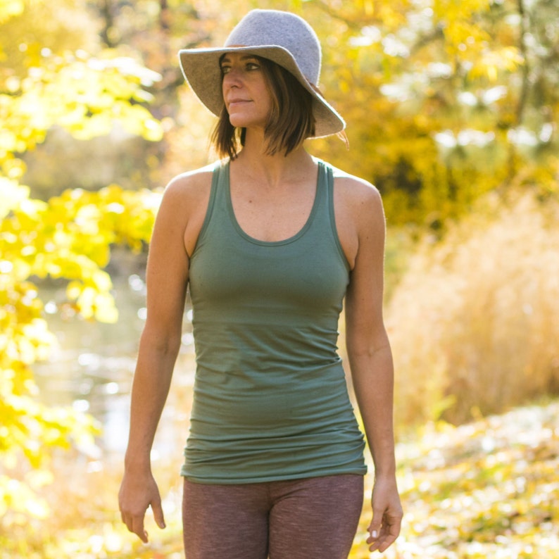 May include: A woman wearing a green tank top and maroon leggings walks through a forest with fall foliage. She is wearing a gray felt fedora.
