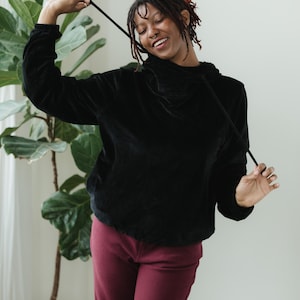 May include: A black velvet hoodie with a drawstring, worn with burgundy corduroy trousers. The outfit is casual. A large green plant in a yellow pot is in the background.