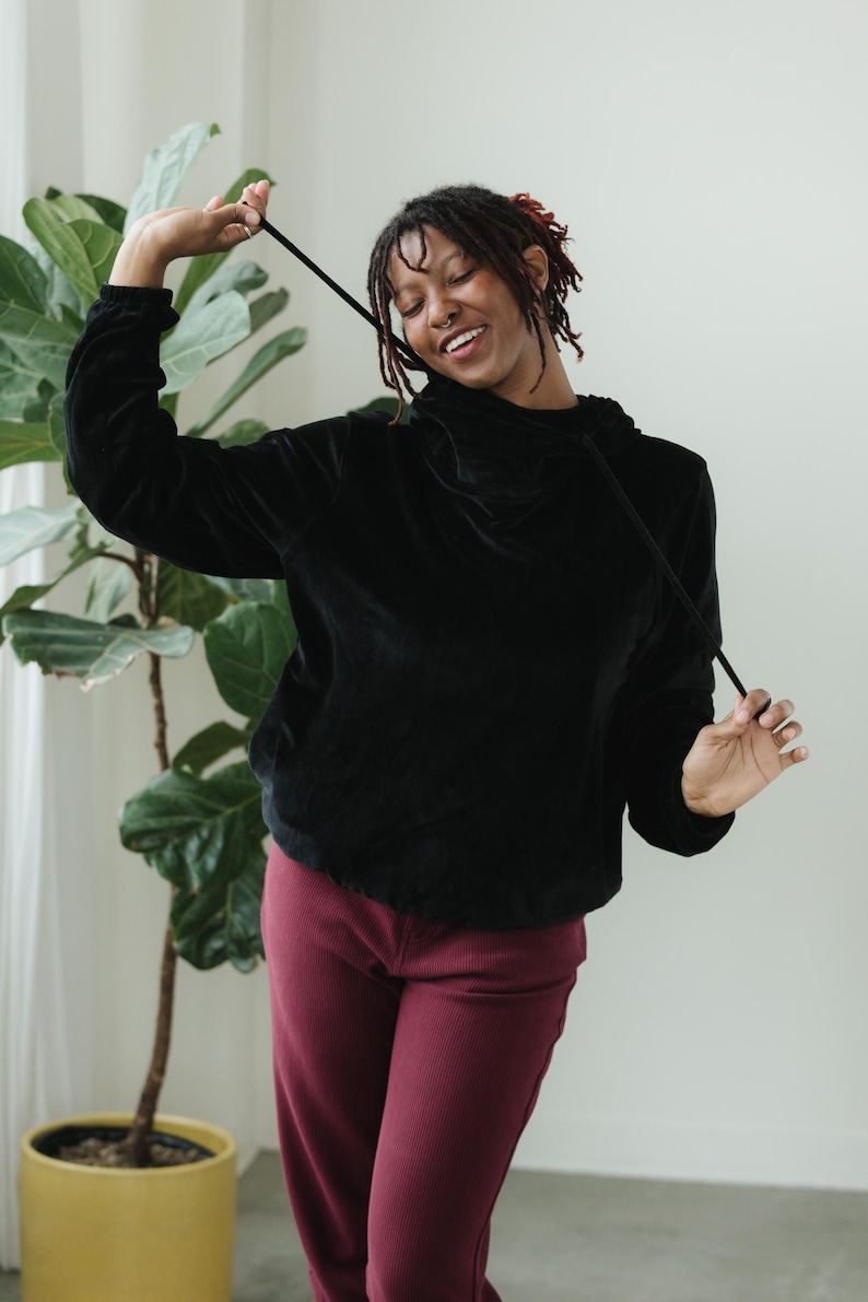 May include: A black velvet hoodie with a drawstring, worn with burgundy corduroy trousers. The outfit is casual. A large green plant in a yellow pot is in the background.