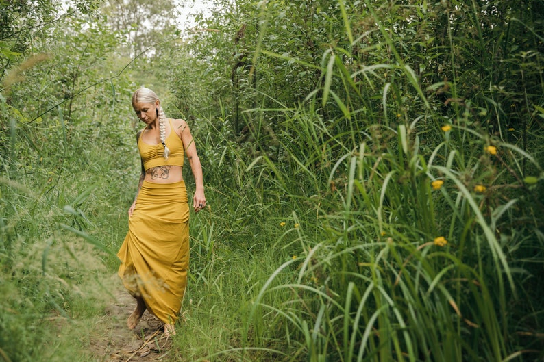 May include: A mustard yellow two-piece set consisting of a cropped tank top with a gathered neckline and a matching floor-length maxi skirt with a wide, fitted waistband. The fabric is a solid, matte mustard yellow color throughout the garment.