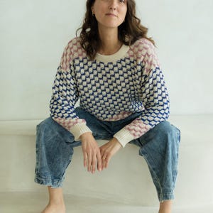 May include: A patterned sweater with a cream-colored base, featuring a repeating design in navy blue and pink. The sweater has a crew neck and long sleeves. The person is wearing blue denim jeans.