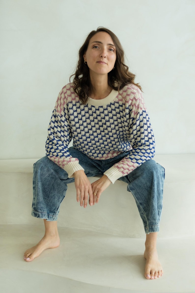 May include: A patterned sweater with a cream-colored base, featuring a repeating design in navy blue and pink. The sweater has a crew neck and long sleeves. The person is wearing blue denim jeans.