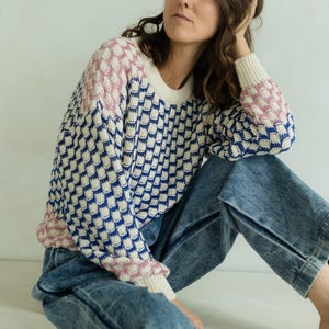 May include: A patterned sweater in cream, navy blue, and pink. The sweater has a crew neck and long sleeves. The model is wearing blue jeans. The sweater's pattern is a repeating geometric design.