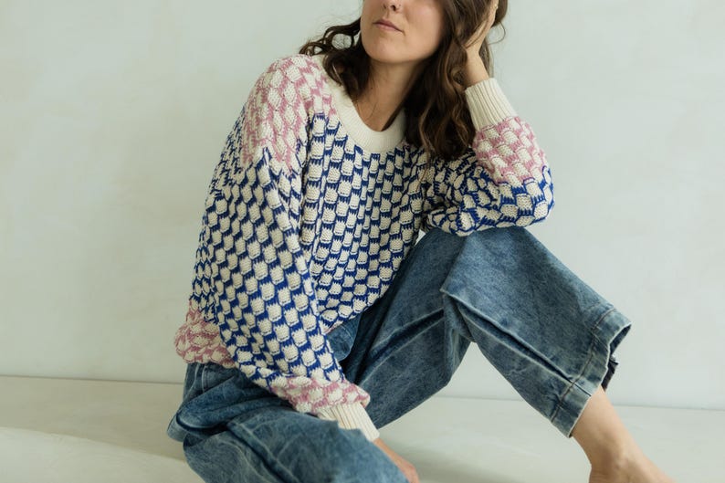 May include: A patterned sweater in cream, navy blue, and pink. The sweater has a crew neck and long sleeves. The model is wearing blue jeans. The sweater's pattern is a repeating geometric design.