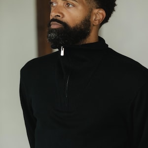 May include: A black long-sleeve shirt with a quarter-zip closure. The shirt has a high collar and a ribbed texture around the zipper. The model has a full beard and is looking to the side.