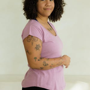 May include: A light purple scoop-neck t-shirt paired with black pants. The person in the image has curly dark hair and tattoos on their arm. The background is a plain, off-white wall.
