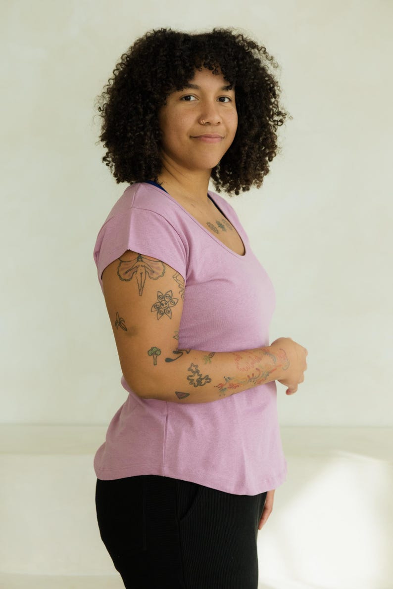 May include: A light purple scoop-neck t-shirt paired with black pants. The person in the image has curly dark hair and tattoos on their arm. The background is a plain, off-white wall.