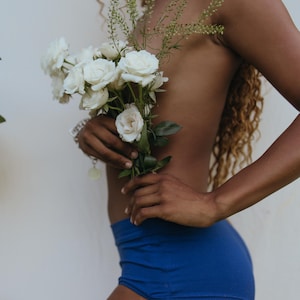 May include: A person holding a bouquet of white roses and greenery, wearing blue shorts. The person's skin is visible, and they are standing in front of a white wall. The image has a natural, soft aesthetic.